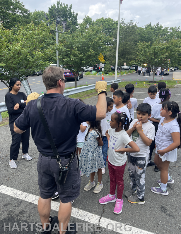 A group of kids listens to a man in a uniform, likely a firefighter, in an outdoor setting.