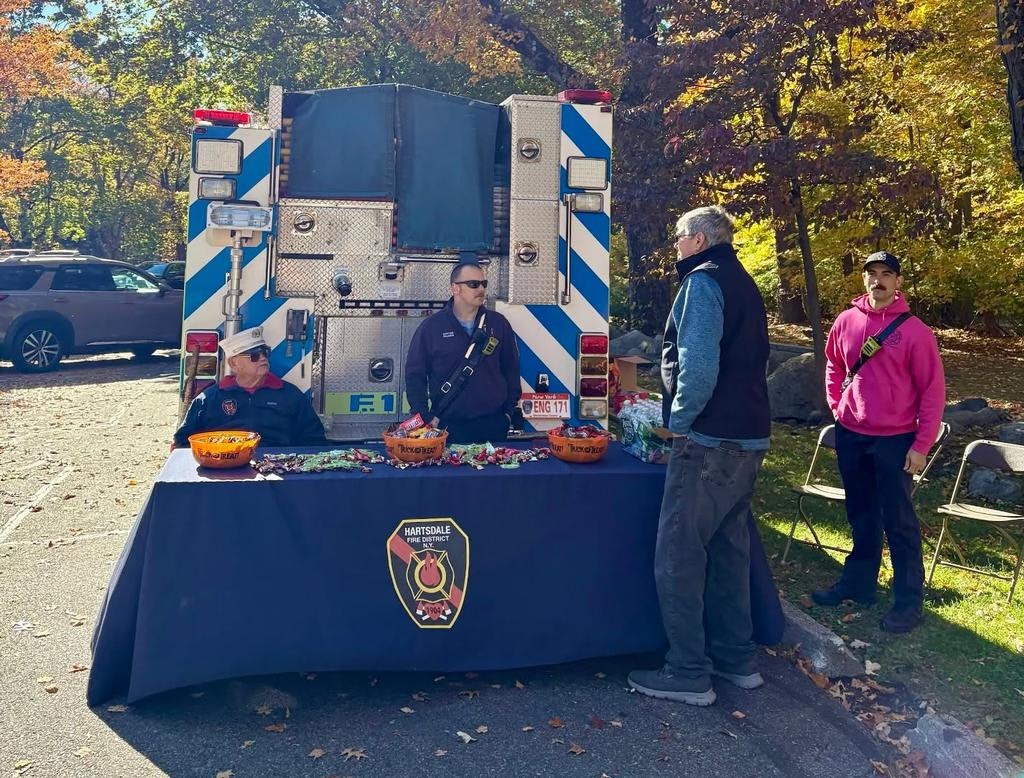 Firefighters at a table offering candy, with a fire truck in the background and fall foliage surrounding the area.