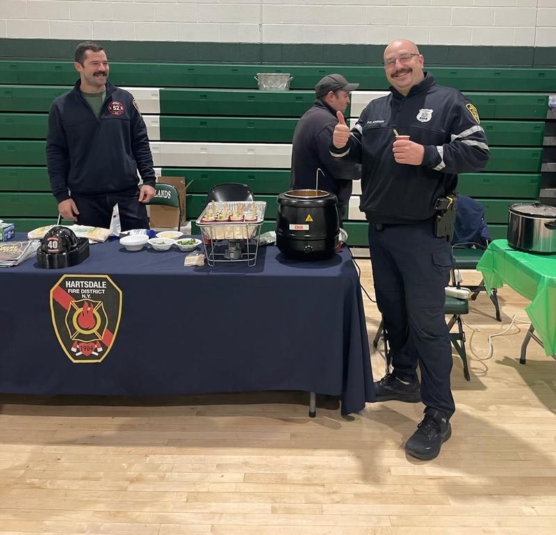 Two men at a table with food, promoting the Hartsdale Fire District at a community event. One man gives a thumbs up.