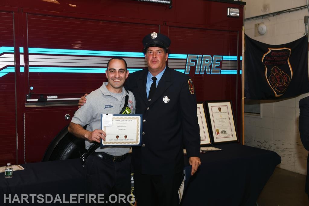 Two firefighters posing with a certificate in front of a fire truck and awards display.