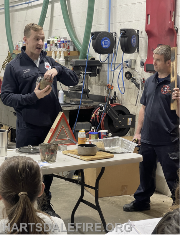 Two firefighters are demonstrating safety equipment and conducting an educational session, with a group observing.