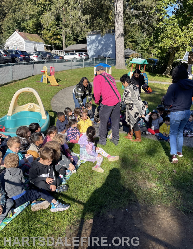 The image shows a group of young children sitting on grass with adults nearby, in a playground setting.