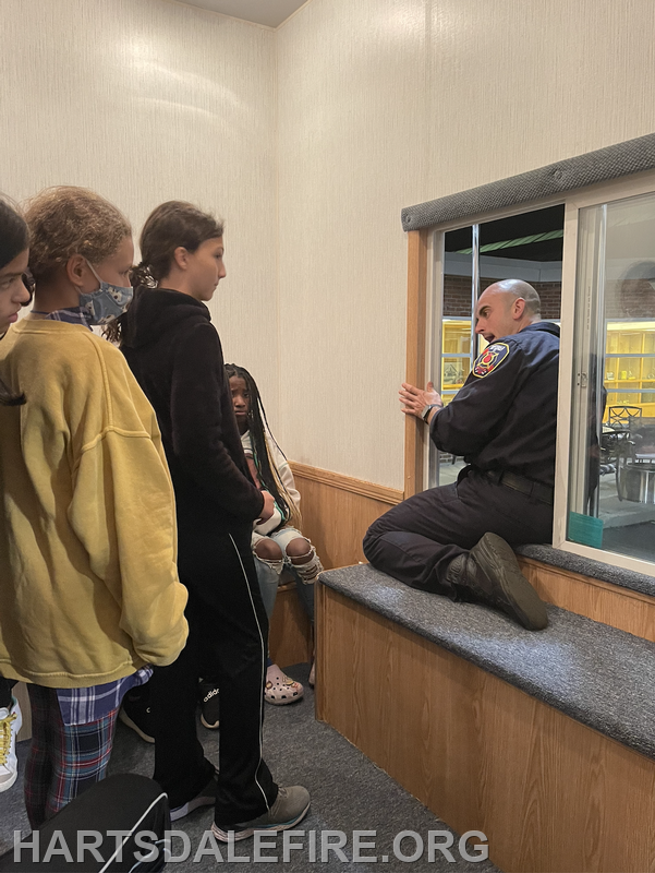 A firefighter talks to attentive kids indoors, sitting by a window.