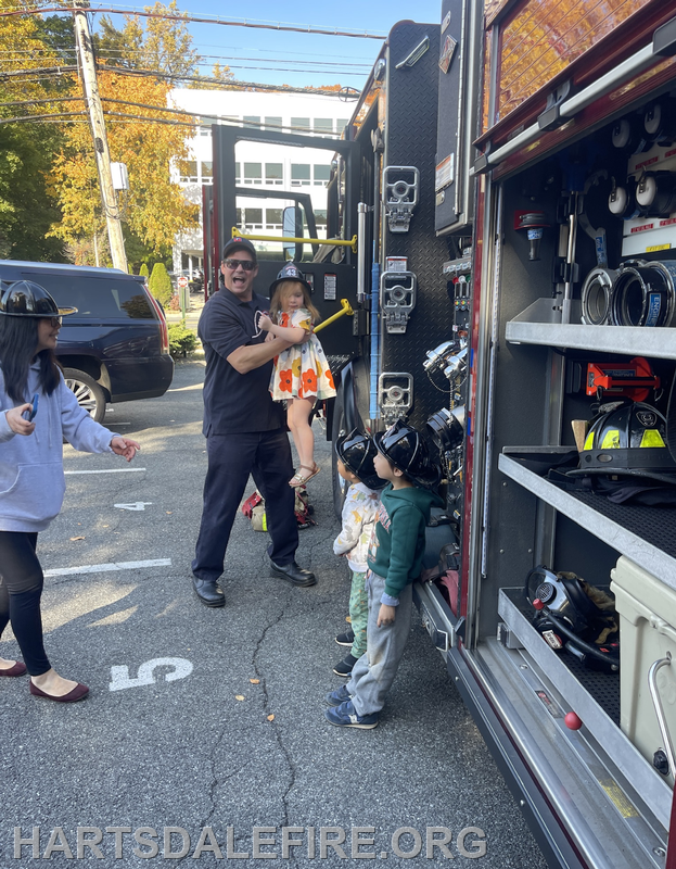 A firefighter engages with children by a fire truck, while a woman looks on. Kids wear helmets and the scene is vibrant.