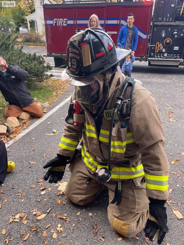 A firefighter in gear kneels on the ground, with a fire truck in the background, while onlookers observe nearby.