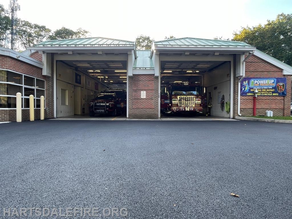 The image shows a fire station with two fire trucks parked inside, a smooth driveway, and a sign promoting fire safety.