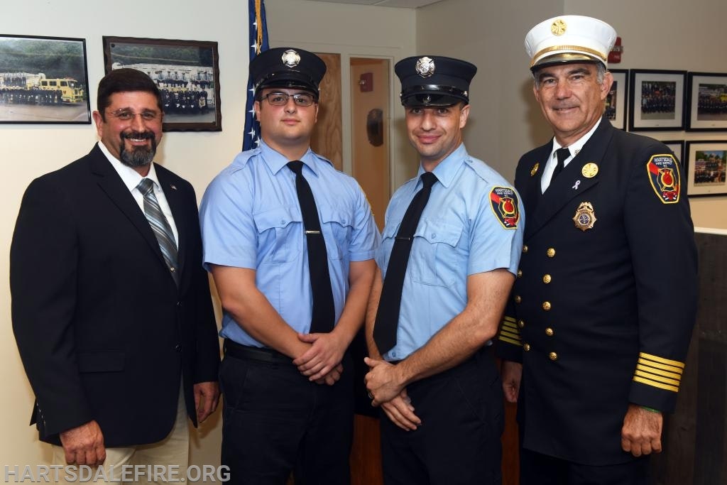 Four men posing; two in light blue uniforms, one in a black uniform with a white cap, and one in a suit, likely at a fire department event.