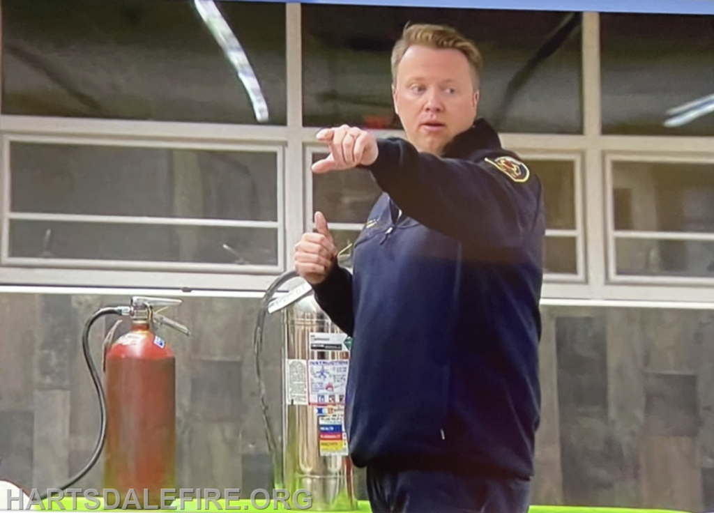 A person in a dark uniform is gesturing while standing near fire extinguishers in a room with windows.