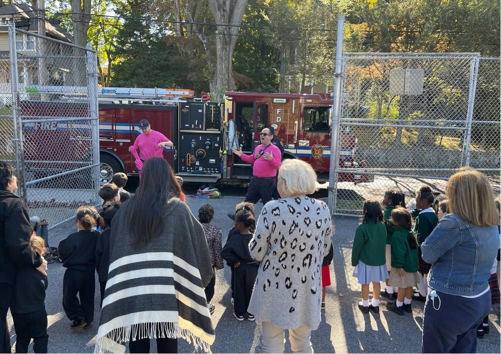 Children gather around a fire truck while firefighters in pink shirts give a presentation.