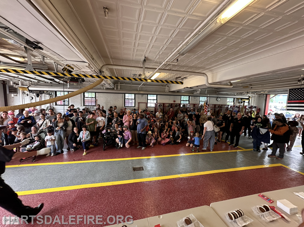 A large crowd gathered inside a fire station, with people of all ages, some holding strollers, and a display of treats on a table.