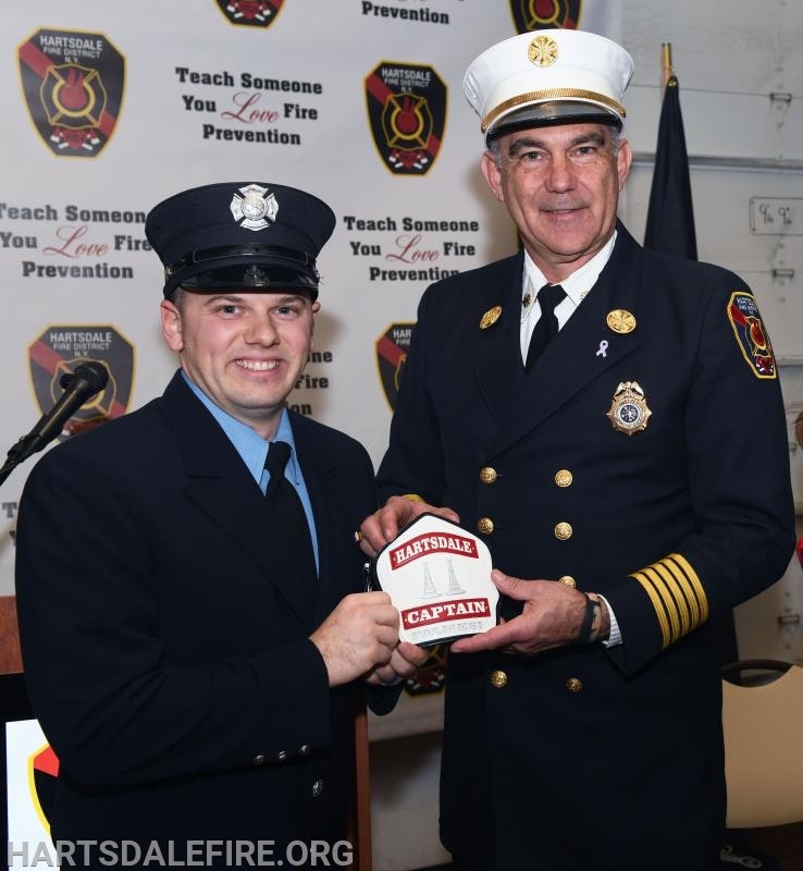 Two firefighters in uniform, holding a "Hartsdale Captain" badge, standing in front of a fire prevention backdrop.