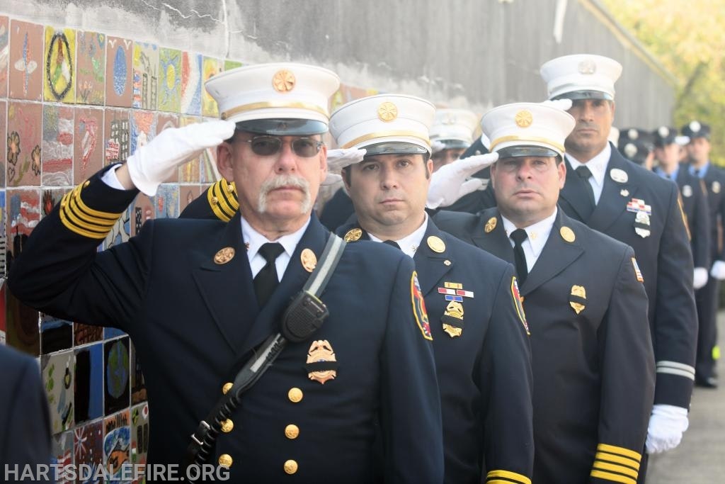 Firefighters in formal uniforms salute, standing in line against a tiled wall.