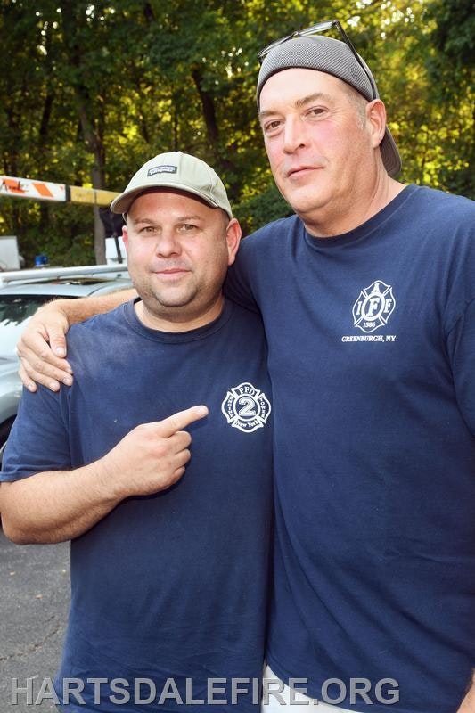 Two men wearing matching blue shirts with firefighter insignias pose together in a friendly manner, outdoors in a wooded area.