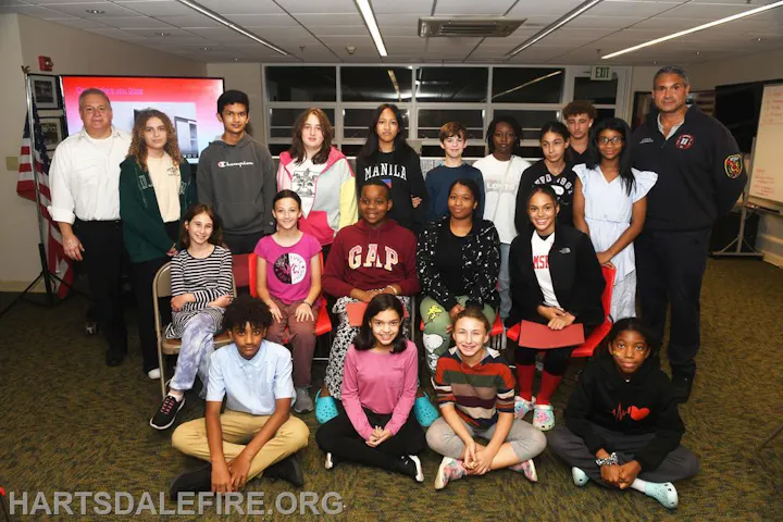 A group of people, including kids and adults, posing for a photo in a room with a screen and whiteboard.