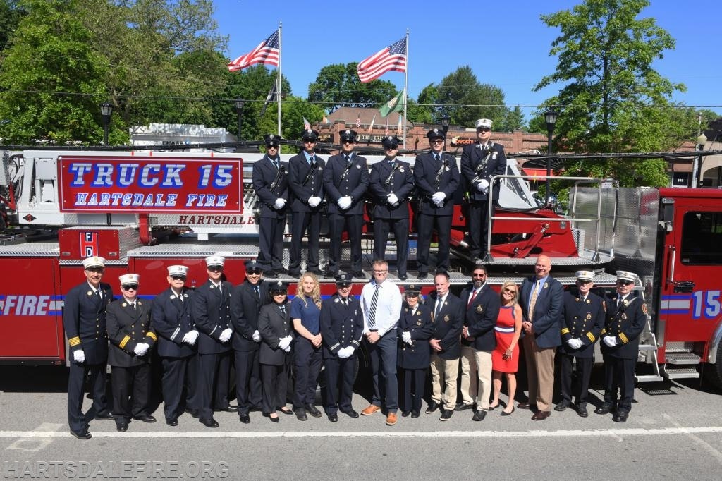 A group of firefighters and officials in formal attire pose in front of a fire truck labeled "Truck 15 Hartsdale Fire."