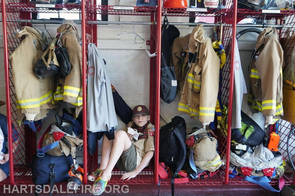 A child wearing a scout uniform is sitting in a fire station locker with firefighter gear hanging above.