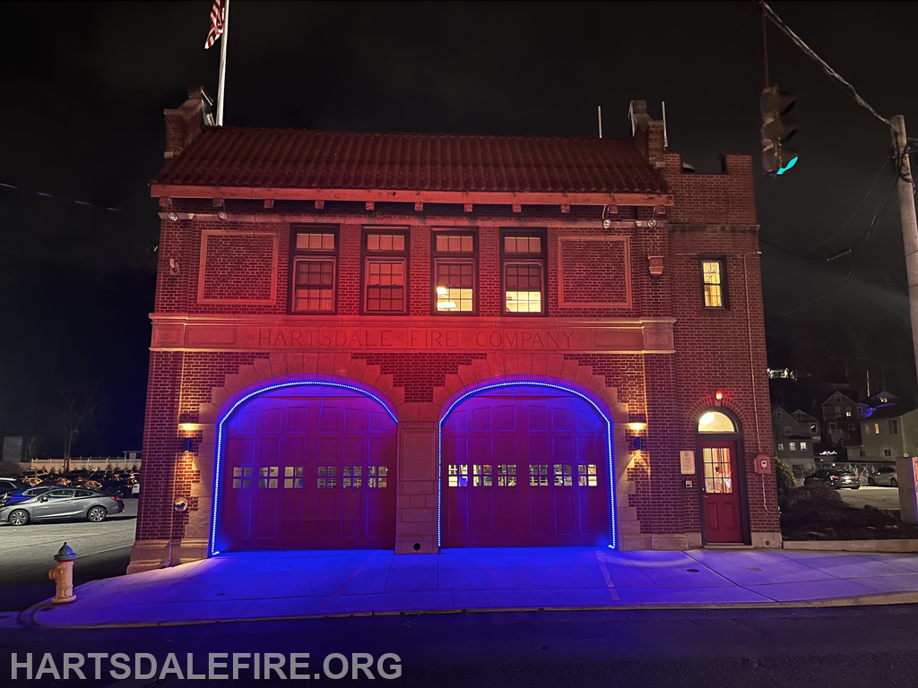 The image shows a night view of the Hartsdale Fire Company building illuminated with red and blue lights.