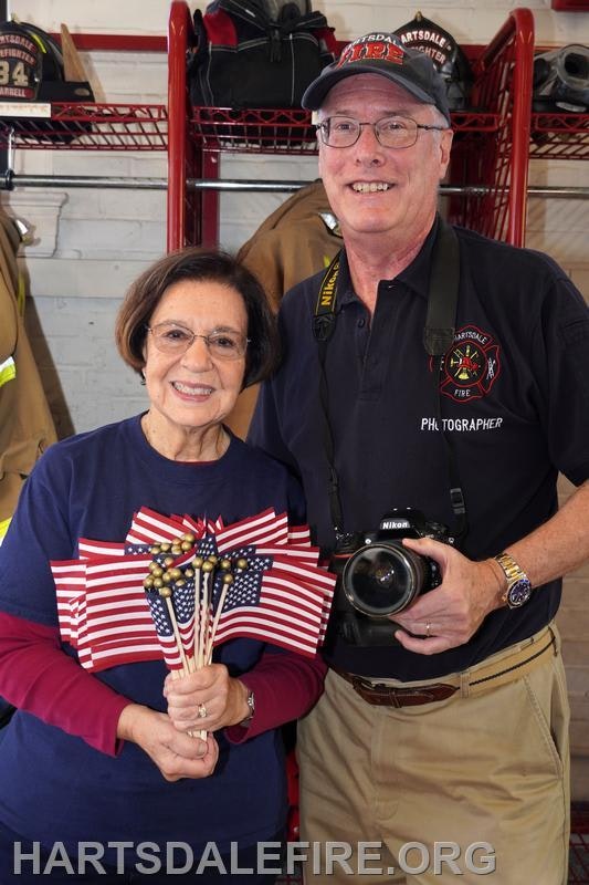 A smiling woman holds small American flags, while a man with a camera poses beside her in a fire station setting.