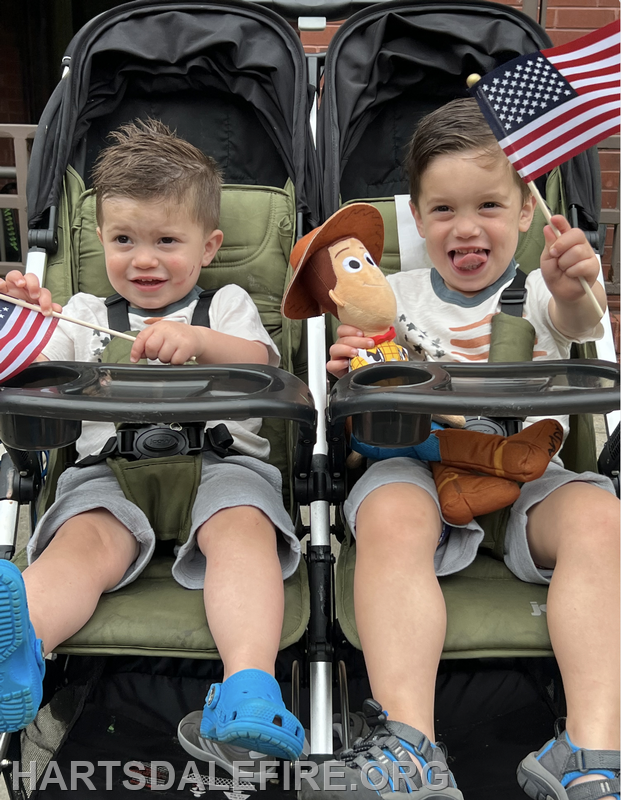 Two young boys in strollers, one holding a toy cowboy and the other a small American flag, both smiling and playful.