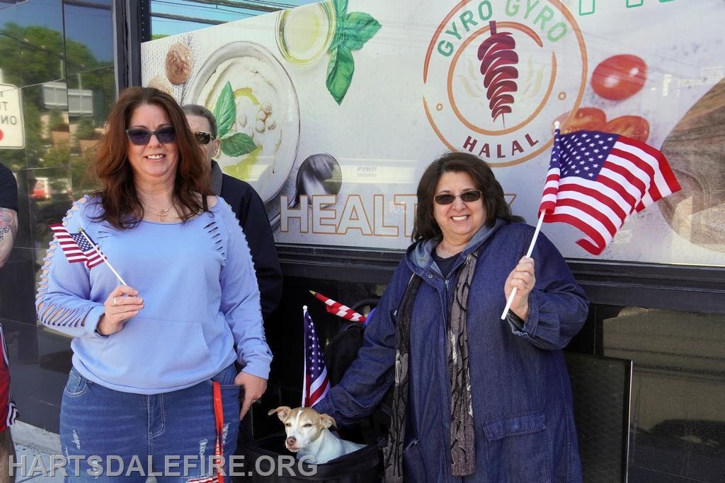 Two women hold American flags outside a restaurant, with a small dog nearby. The setting appears festive and community-oriented.