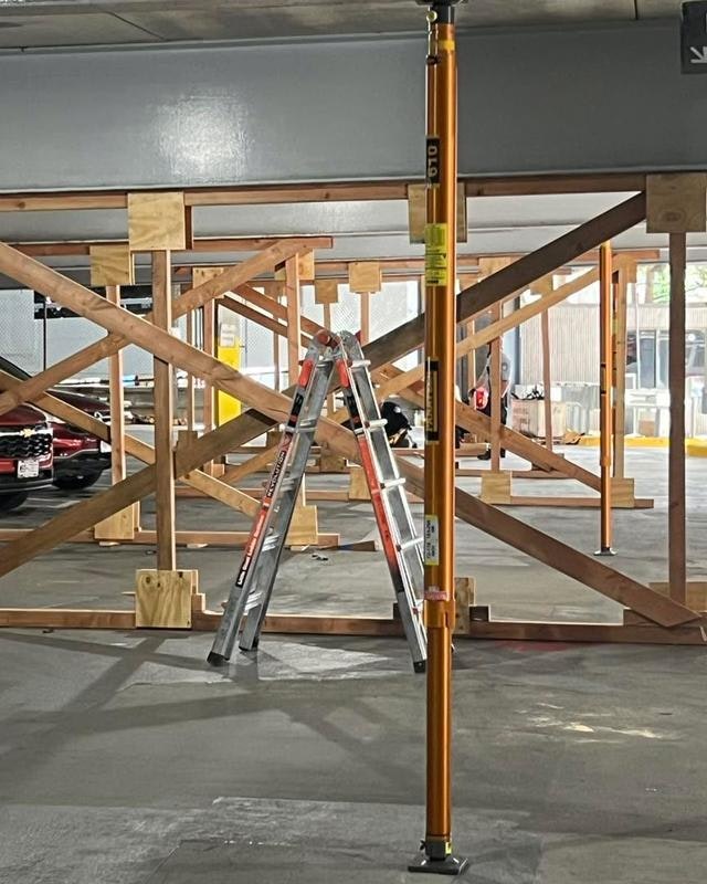 The image shows a parking area under construction, featuring wooden supports, a ladder, and a support pole.