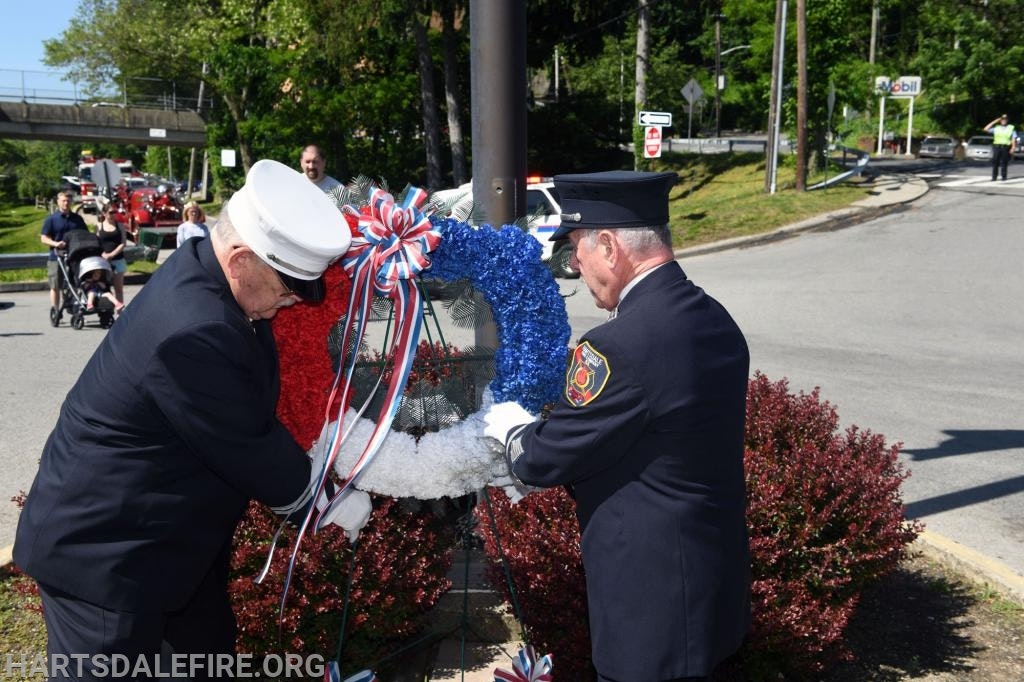 Two firefighters in uniform place a red, white, and blue wreath at a memorial.