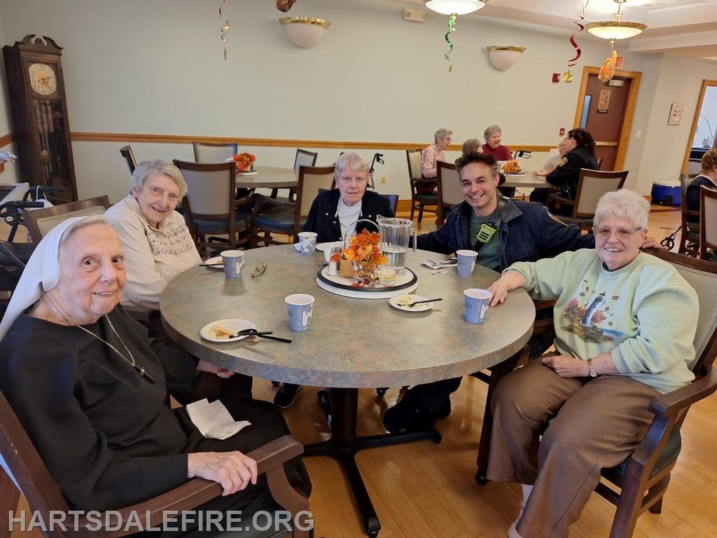 Five people sit around a table enjoying a meal, with festive decorations in the background, in a cheerful communal setting.
