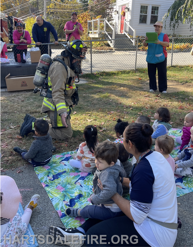A firefighter interacts with young children during a community event, while others observe nearby.