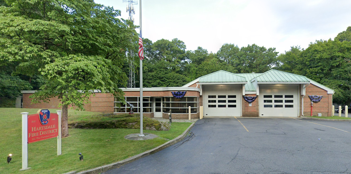The image shows the Hartsdale Fire District building, featuring a red sign, garage doors, and greenery surrounding the area.