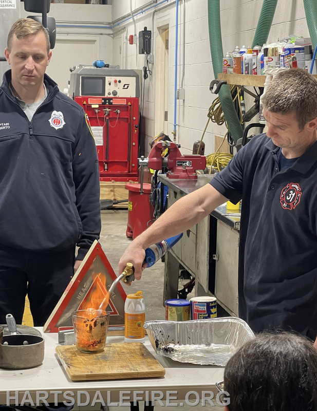 A firefighter demonstrates a flame in a workshop, while another looks on, highlighting safety and fire techniques.