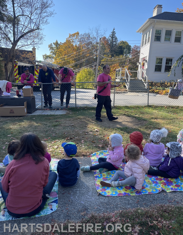 Firefighters in pink shirts engage with children sitting on blankets, likely at a community event, with a sunny backdrop.