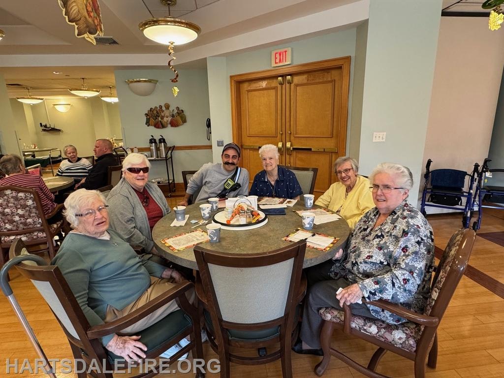 A group of six people, mainly seniors, gathers around a table with snacks in a communal space. They appear to be socializing and enjoying each other's company.