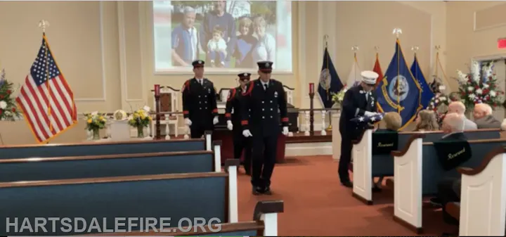 A solemn ceremony with firefighters in uniform, an American flag, and attendees seated, likely honoring a fallen comrade.