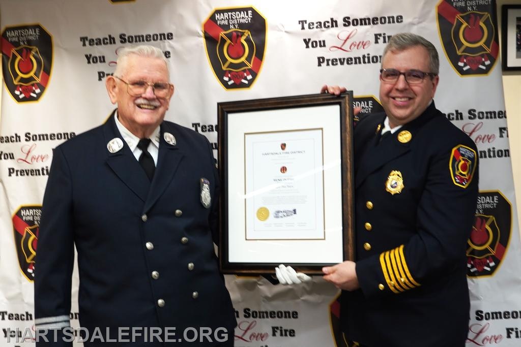 Two men in fire service uniforms are holding a framed certificate, standing in front of a backdrop promoting fire prevention.