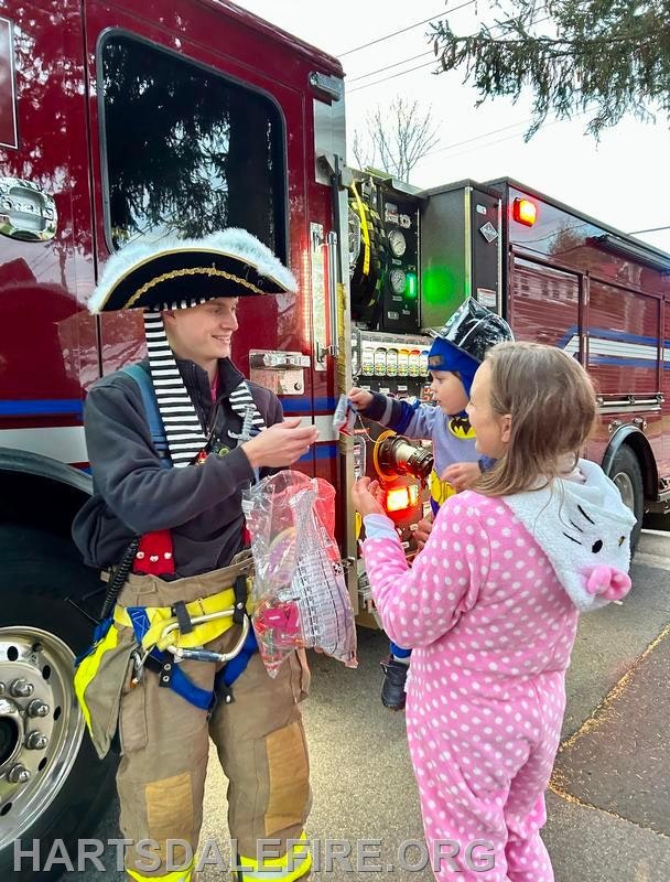 A firefighter in a pirate hat hands treats to two children in costumes near a fire truck. Fun and festive atmosphere!
