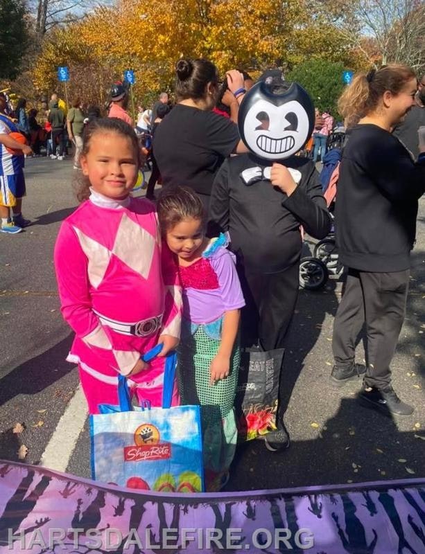 Three kids in costumes pose for a photo at a festive outdoor event, surrounded by people and autumn foliage.
