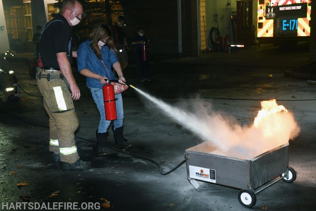 A firefighter instructs a person using a fire extinguisher on a small controlled fire outdoors.
