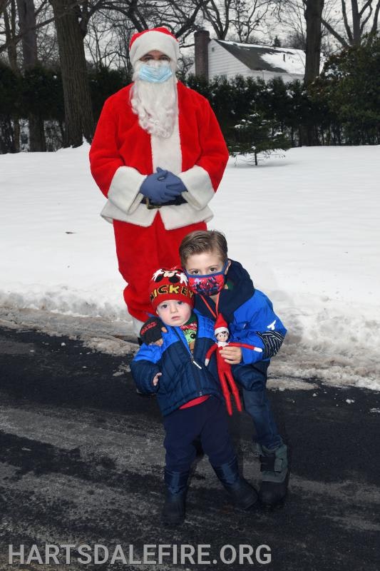 Two children in winter clothes stand in front of a Santa Claus figure wearing a mask and gloves, outdoors in the snow.