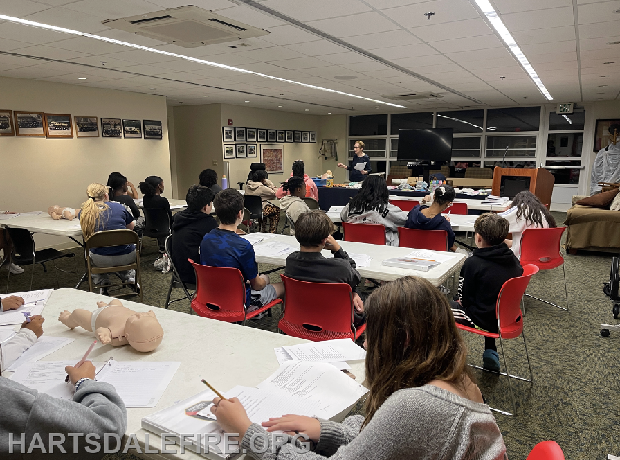 A group of students attends a classroom session, learning with a presentation and using practice mannequins for exercises.