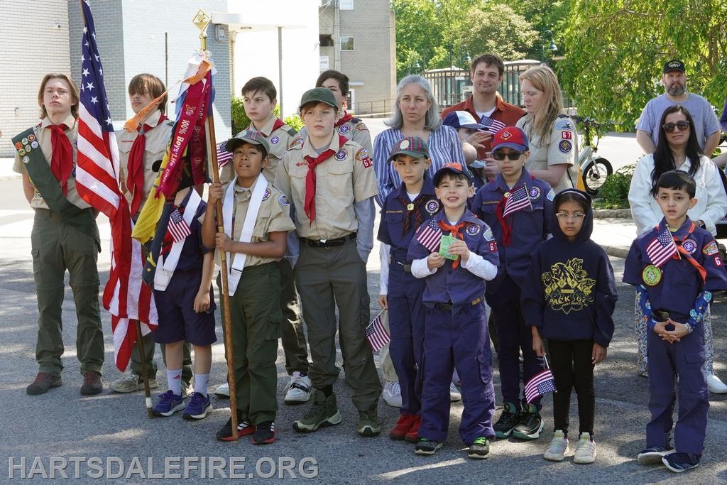 A group of children and adults, some in uniforms, hold flags and stand together outdoors, likely at a community event.