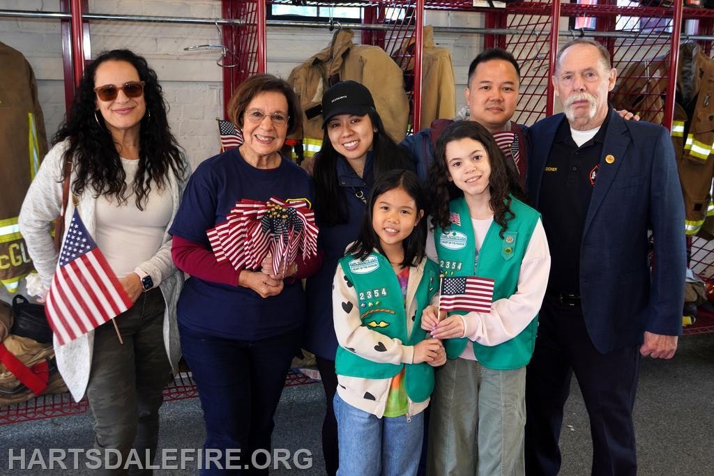 A group of individuals, including children and adults, are posing together with American flags in front of firefighter gear.