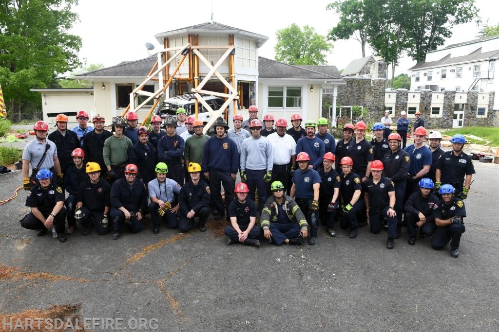 A group of firefighters in helmets posing for a photo in front of a house with structural supports.