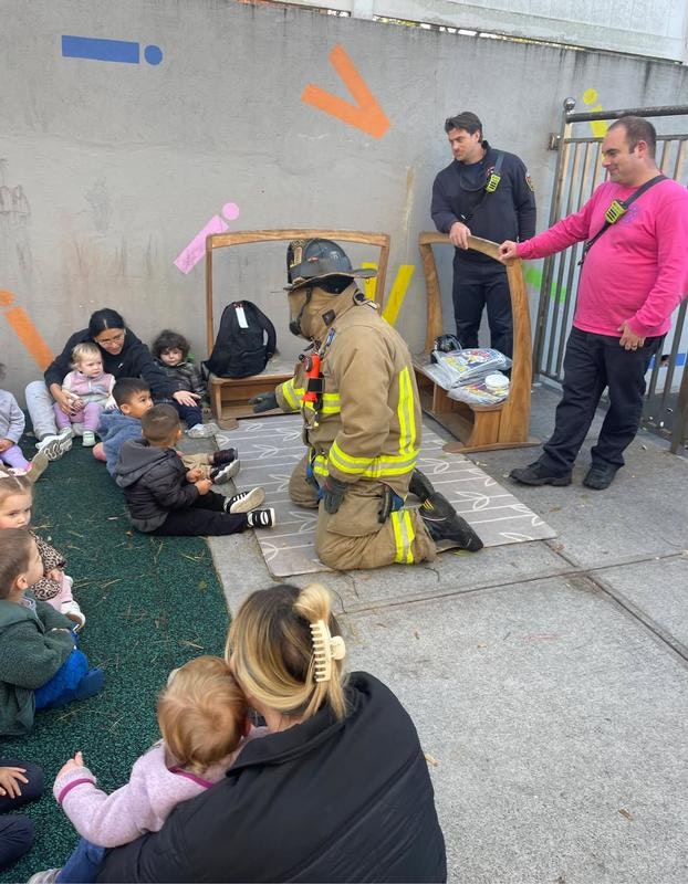 A firefighter interacts with a group of children sitting on a mat, while adults observe in a colorful outdoor setting.