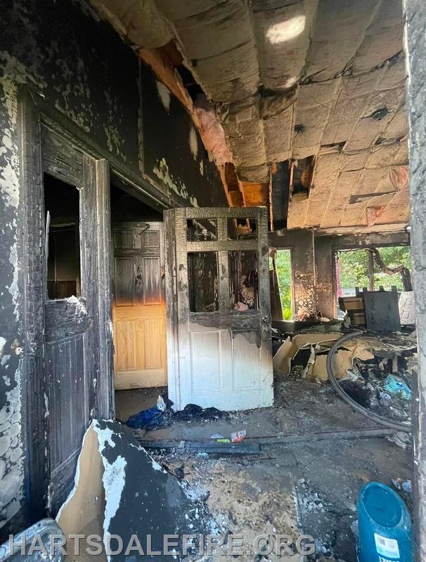 Interior of a severely fire-damaged room with charred walls, doors, and ceiling. Debris is scattered on the floor.
