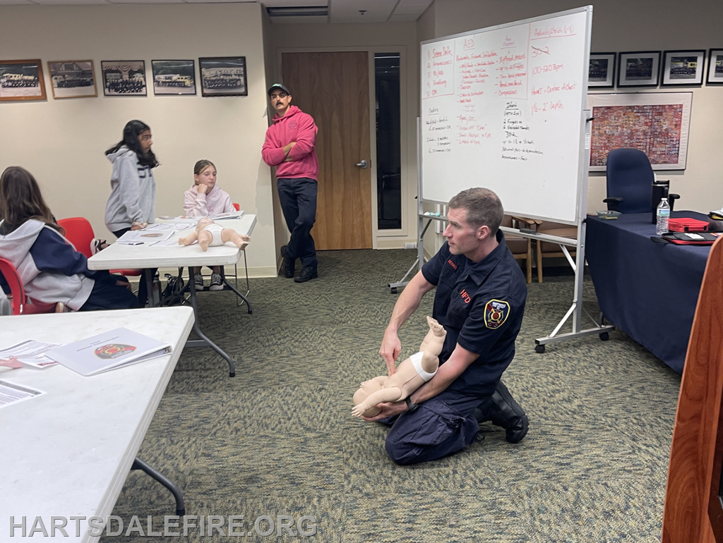 A firefighter demonstrates CPR on a mannequin while students observe in a classroom setting.