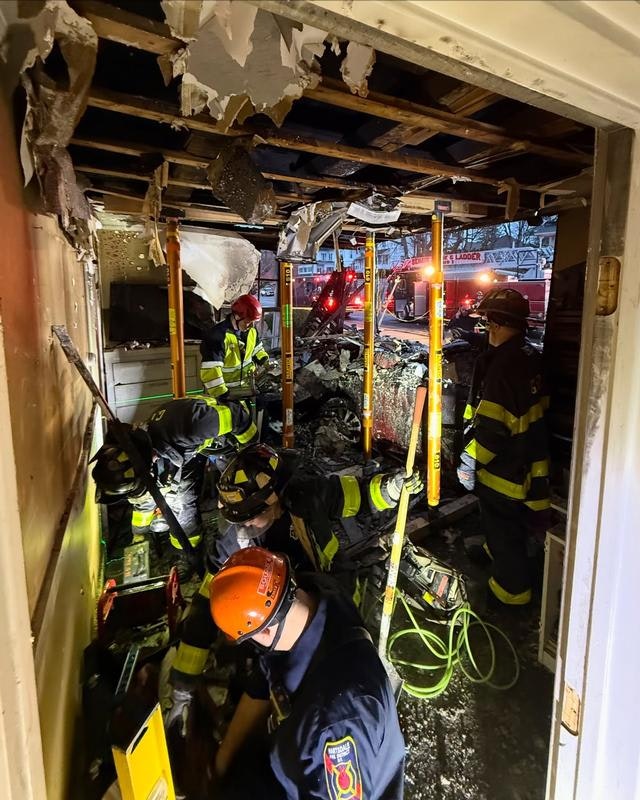 Firefighters are working in a damaged indoor area, removing debris and assessing damage after a fire. Emergency vehicles are visible outside.