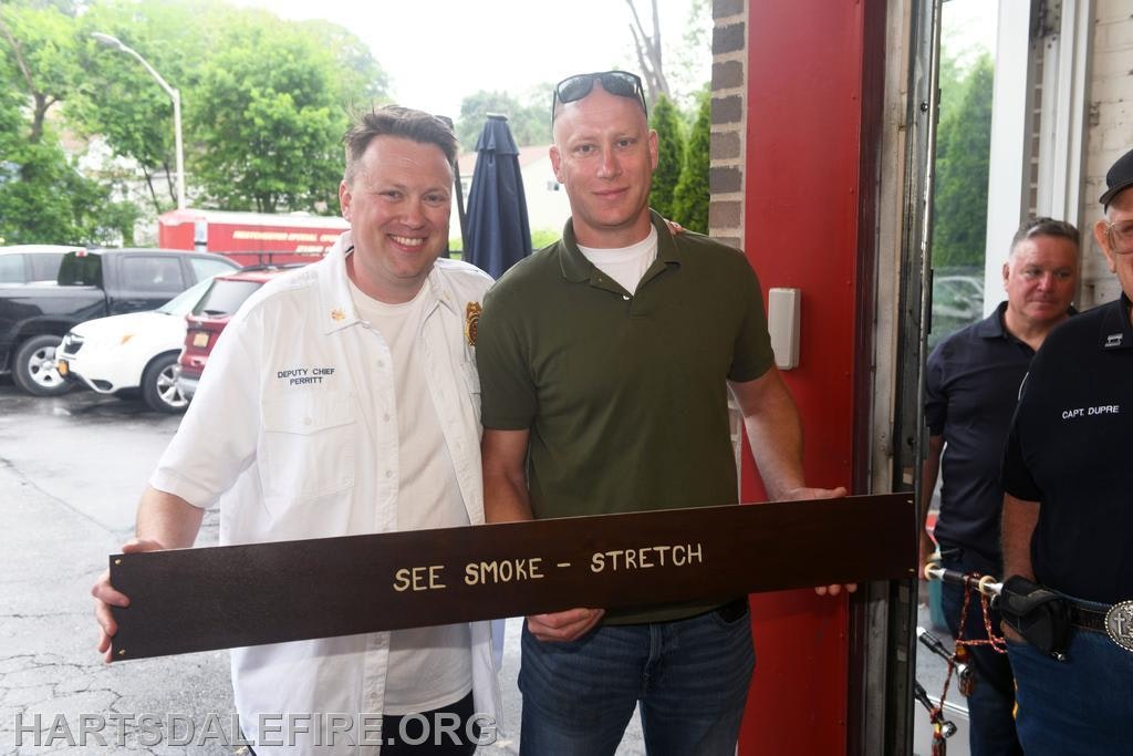 Two men pose with a sign reading "SEE SMOKE - STRETCH" at a fire department event, while others look on in the background.