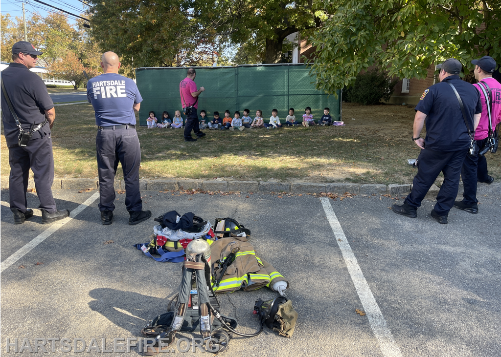 Firefighters are interacting with a group of children outdoors, showcasing equipment while teaching fire safety.