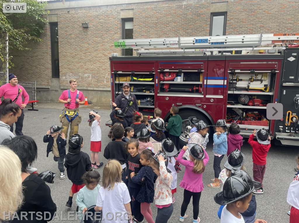 Kids wearing toy fire helmets gather around firefighters by a fire truck, learning about fire safety and equipment.