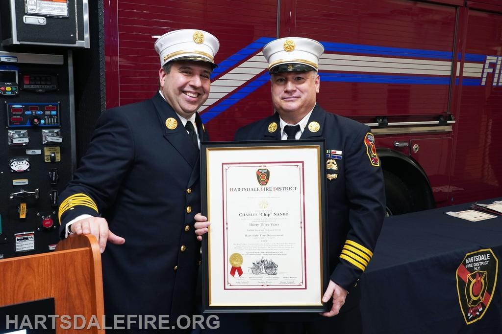 Two firefighters in uniform, one holding a framed certificate, standing in front of a fire truck.